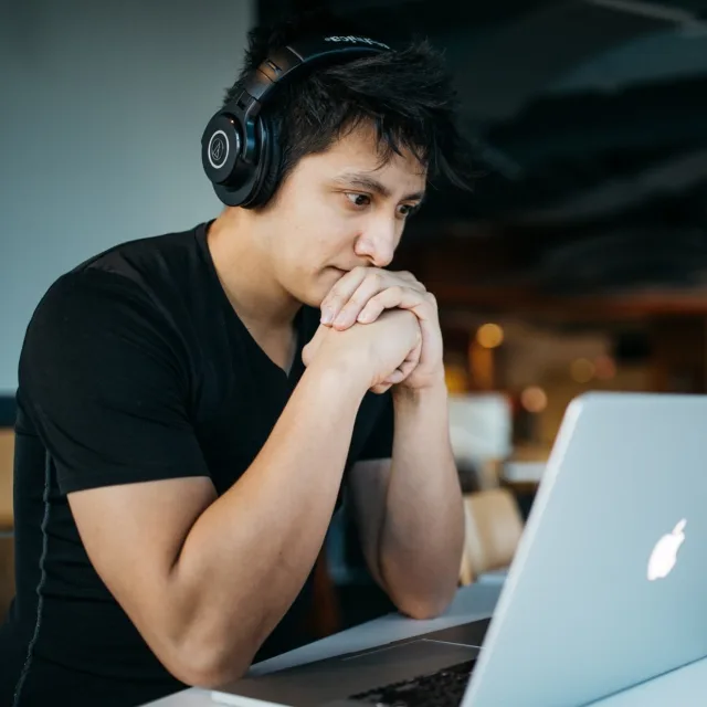 Young man with headphones on, forehead resting on his hands, tense look at his laptop