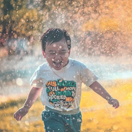 A little boy runs laughing through drops of water, in late-afternoon light
